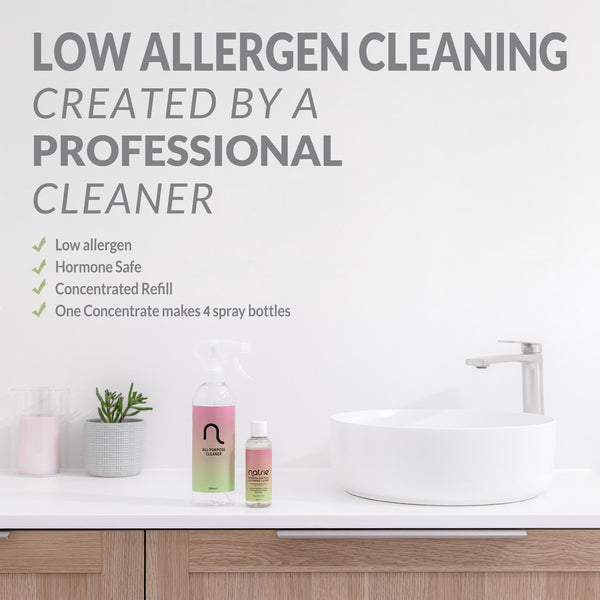 A wall-mounted text graphic promotes low allergen cleaning with a list of benefits and four spray bottles displayed on a bathroom counter beside a white sink and faucet.